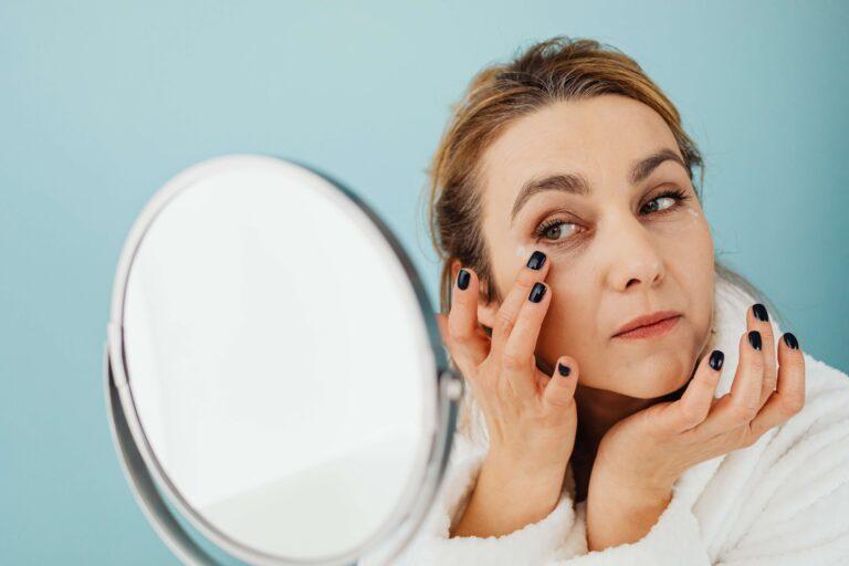 White Woman looking at herself in the mirror while touching under her eye, analyzing her complexion