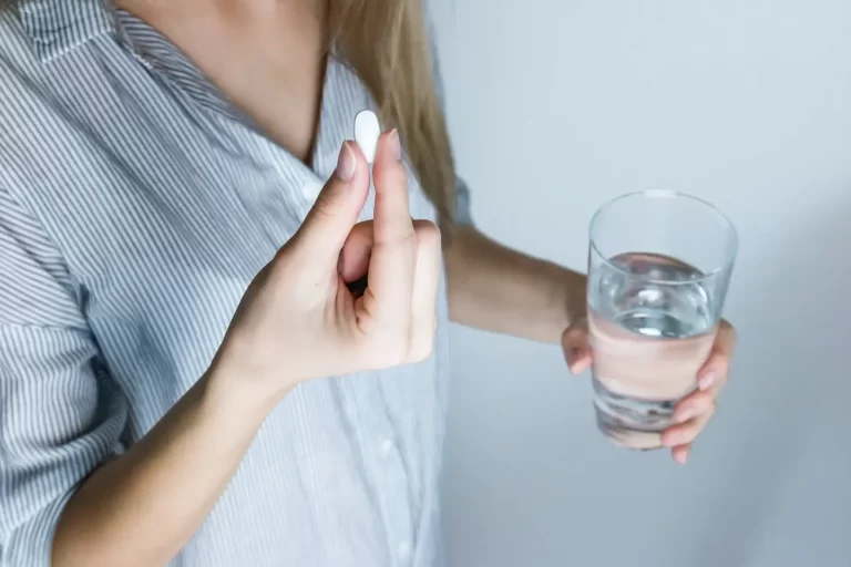 White woman in a light blue button down shirt holding a white gut health supplement and a glass of water. Gut Halth Supplements