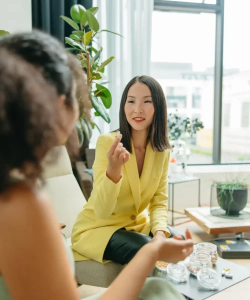 Asian Woman in yellow business suite jacket holding a prebiotic vitamin, offering it to a black woman in green dress. Are prebiotics good for you?