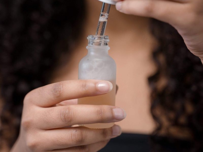 Black woman with long hair holding a clear glass bottle with the cap in her hand. Benefits of Glycolic acid for dandruff